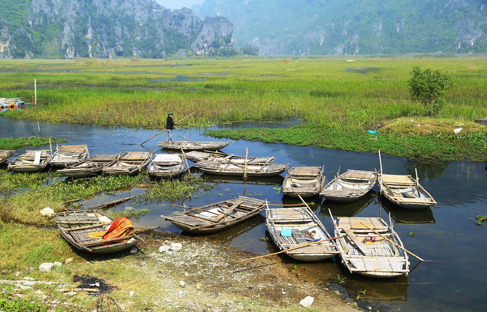 Wooden rowboats moored on a reedy waterway beneath limestone karsts in Ninh Binh's Van Long wetland.
