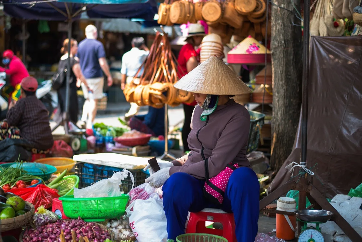 A woman in a conical hat sits selling vegetables at a busy street market in Hoi An.