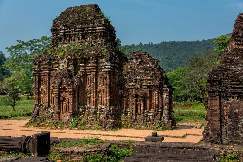 Ancient brick towers of the My Son sanctuary stand amid greenery under a clear blue sky.