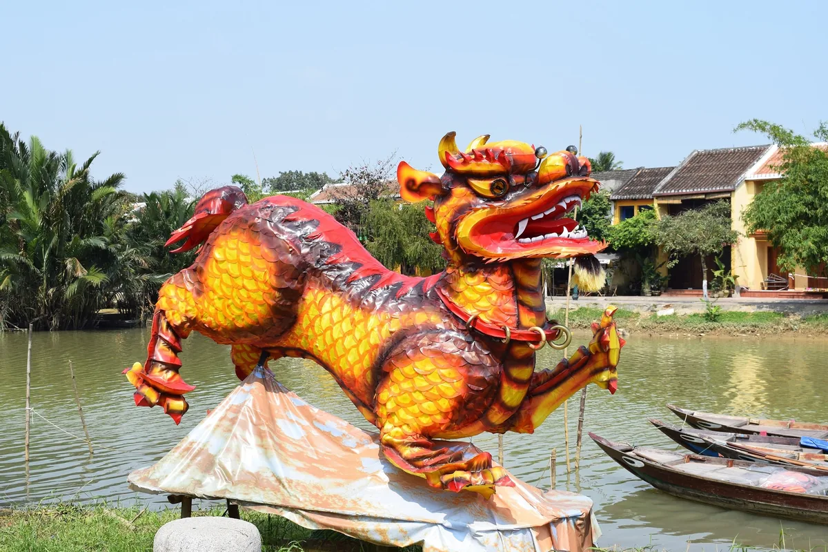 A colorful dragon lantern statue stands beside the Thu Bon River in Hoi An, with wooden boats and old houses in the.