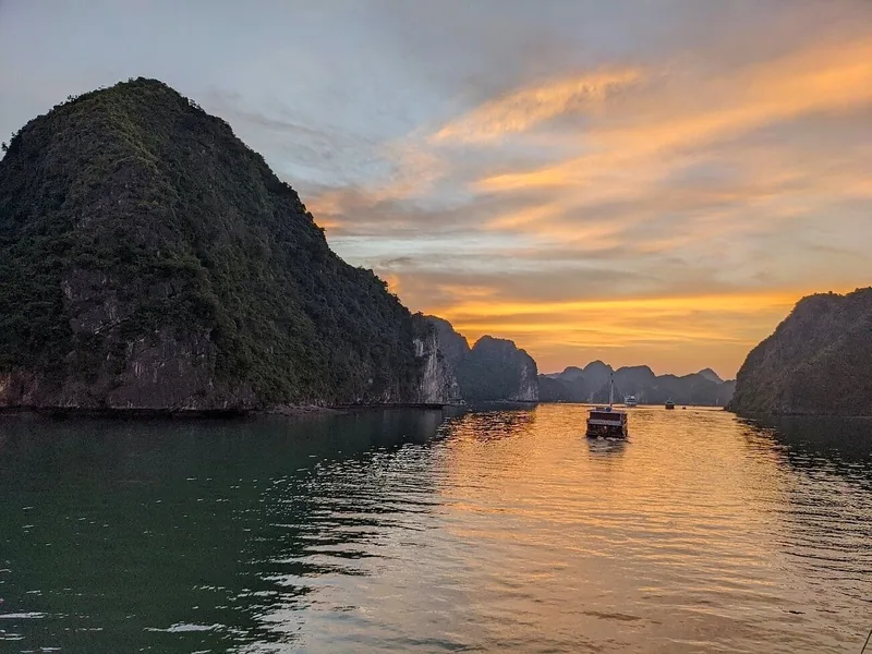 A cruise boat drifts among limestone karsts as the sky glows orange at sunset in Lan Ha Bay.