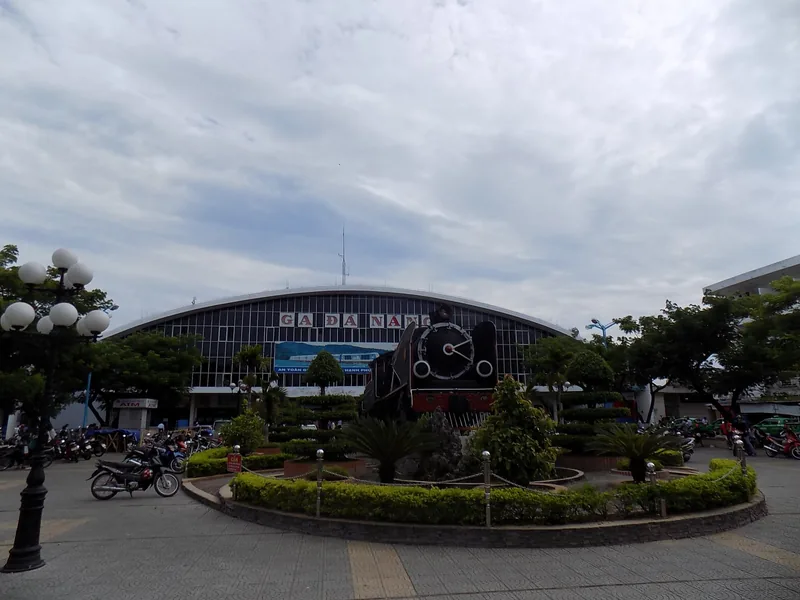 Old steam locomotive display in front of the arched facade of Da Nang railway station under a cloudy sky.