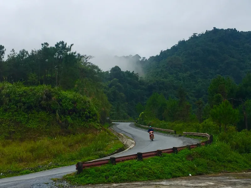A motorbike rides a winding mountain road through misty forested hills in Ha Giang, Vietnam.