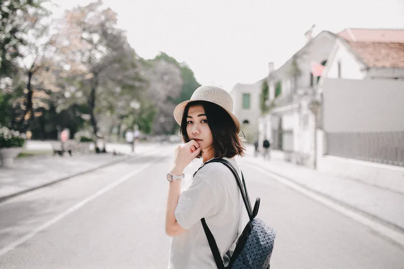 A young woman in a straw hat and backpack pauses on a quiet tree-lined street in Hanoi.