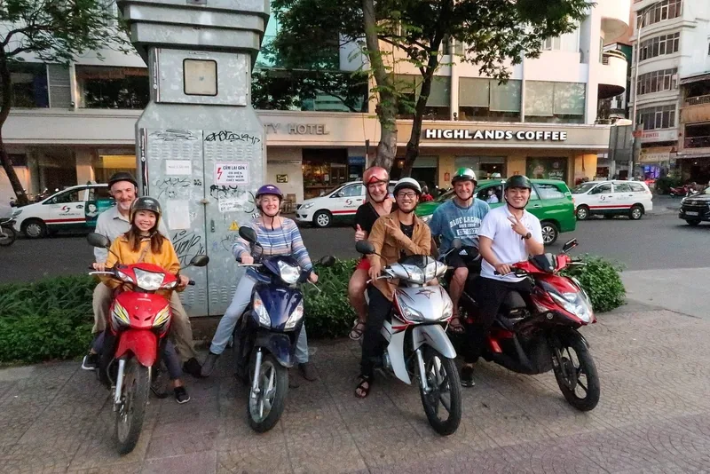 Tourists on motorbikes with local guides pose on a Saigon street near Highlands Coffee at dusk.