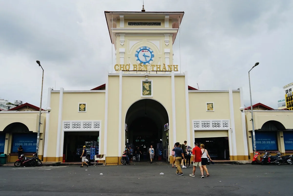 The iconic clock tower entrance of Ben Thanh Market in Saigon with visitors walking past on an overcast day.