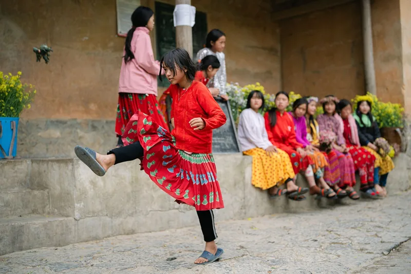 A girl in colorful traditional dress performs a dance step while others watch from a stone wall in Ha Giang.