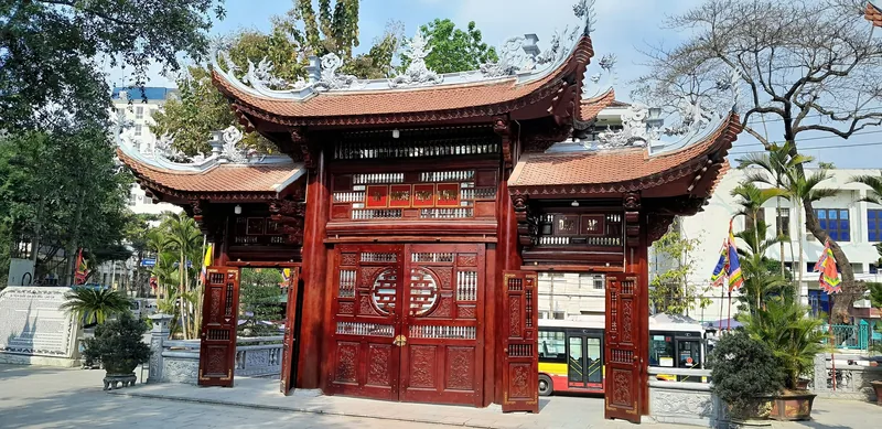 An ornate red wooden gate with traditional dragon carvings and curved tiled roof stands at a temple entrance in Lao Cai city.