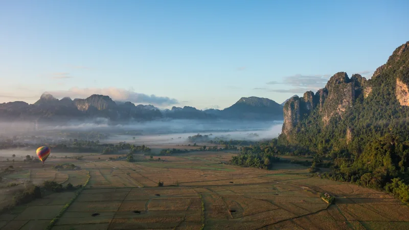 A hot air balloon drifts over misty karst mountains and rice fields at dawn in Vang Vieng, Laos.