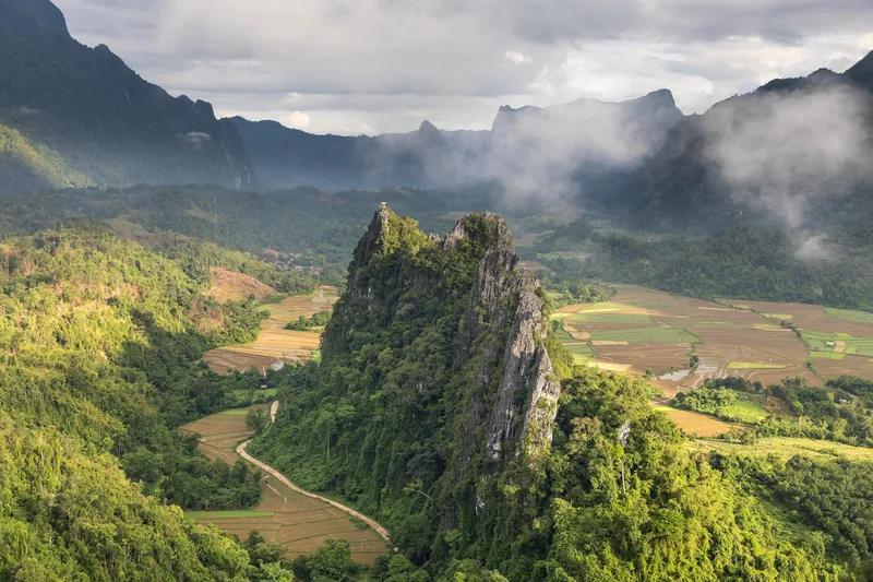 A jagged limestone karst rises above green rice fields and misty mountains near Vang Vieng, Laos.