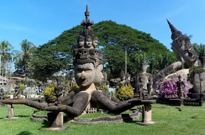 Multi-headed concrete statue and other Buddhist sculptures in Buddha Park, Vientiane, under a clear blue sky.
