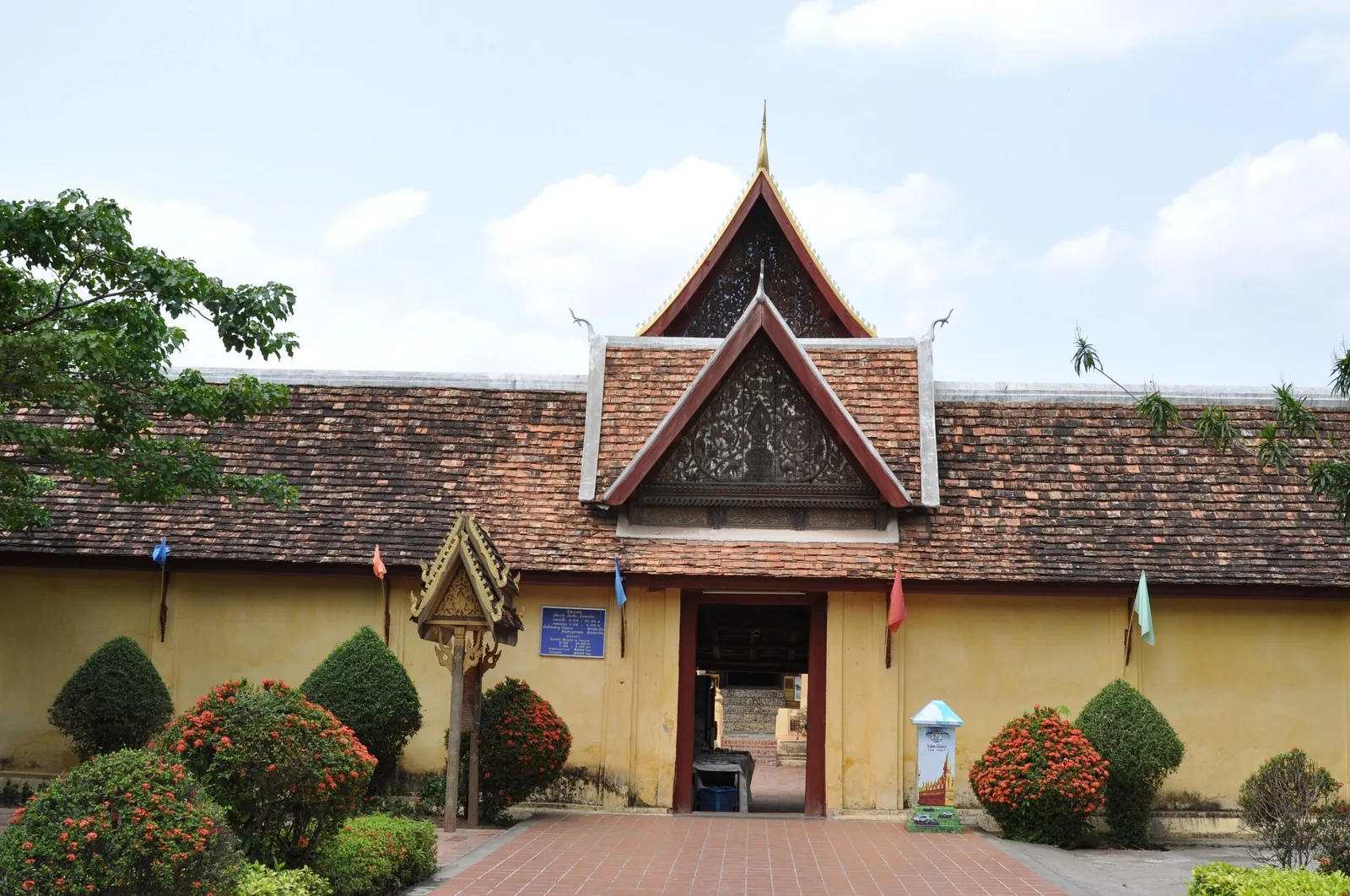 Ornate tiled gable roof over the entrance gate of a temple compound in Vientiane, framed by trimmed shrubs.