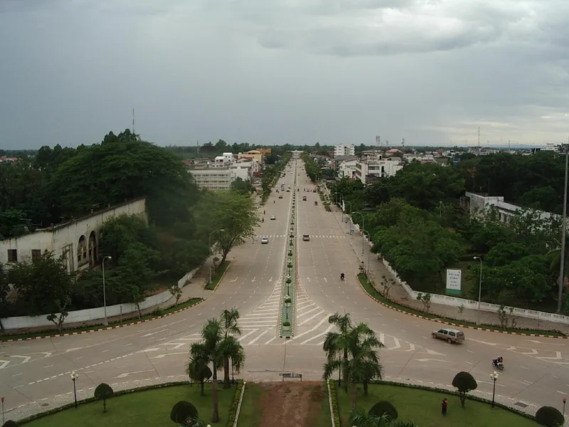 Aerial view of a wide boulevard lined with trees and buildings stretching into the city of Vientiane under a cloudy sky.