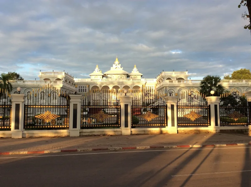 A grand white palace building with golden decorative gates stands behind an ornate fence in Vientiane.
