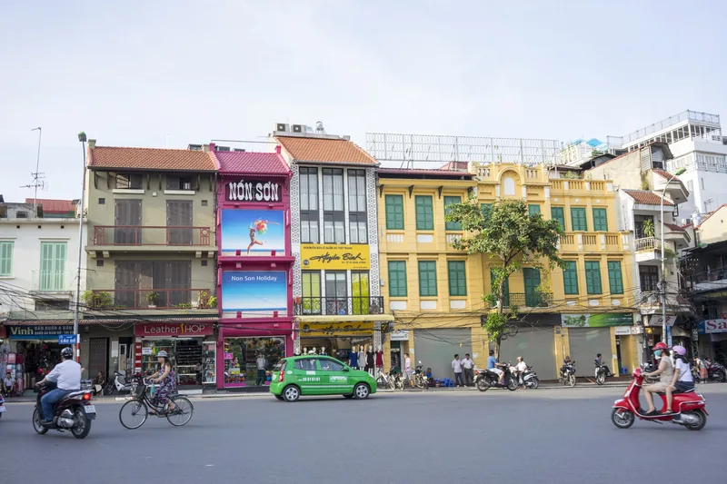 Colorful shophouses line a busy street in Hanoi's Old Quarter with motorbikes and a taxi passing by.