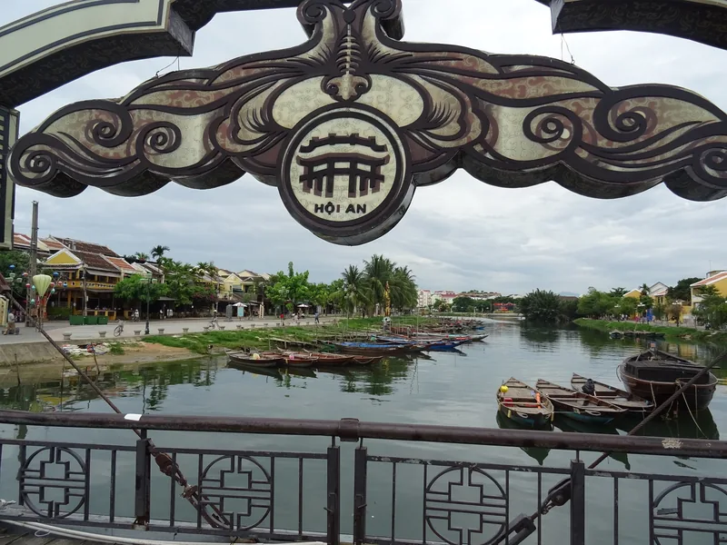 An ornate Hoi An sign arches over the Thu Bon River, with wooden boats moored along the water below.