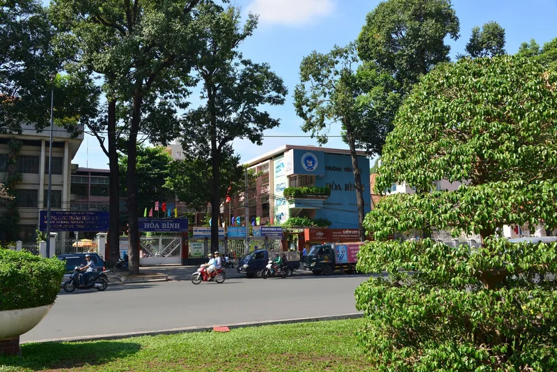 A tree-lined street in Ho Chi Minh City with motorbikes passing the Hoa Binh school building.