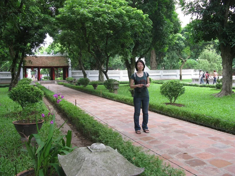 A woman stands on a brick path lined with hedges and trees within the Temple of Literature grounds in Hanoi.