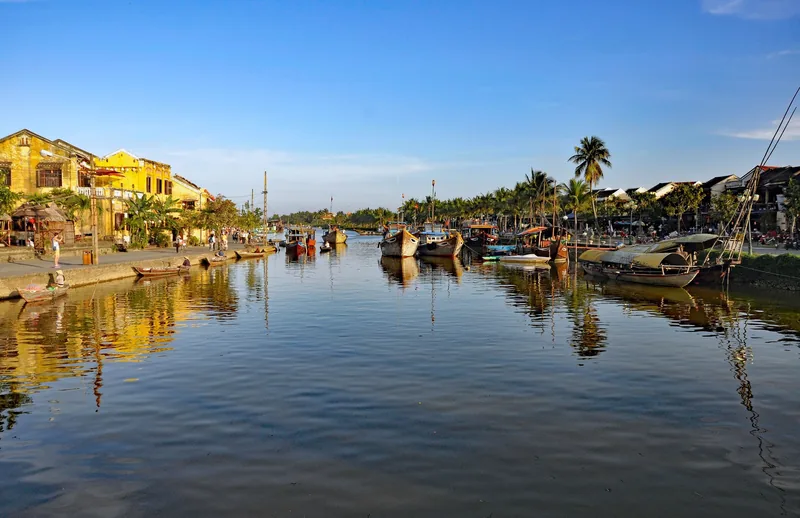 Wooden boats line the calm Thu Bon River beside Hoi An's ancient yellow-walled houses at golden hour.