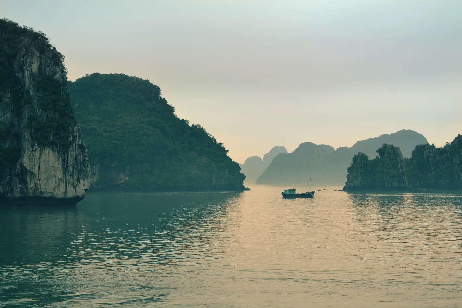 A small fishing boat drifts among misty limestone karsts at dawn in Ha Long Bay, Vietnam.