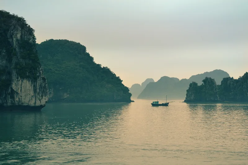 A small fishing boat drifts among misty limestone karsts at dawn in Ha Long Bay, Vietnam.