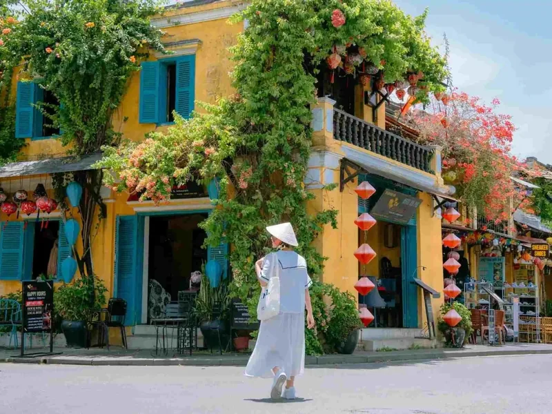 A woman in a conical hat walks past a yellow, vine-covered building strung with red lanterns in Hoi An.