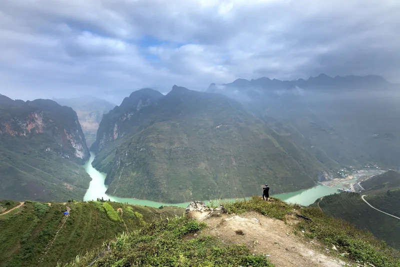 A person stands on a cliff overlooking the turquoise Nho Que River winding through steep limestone mountains in Ha Giang.