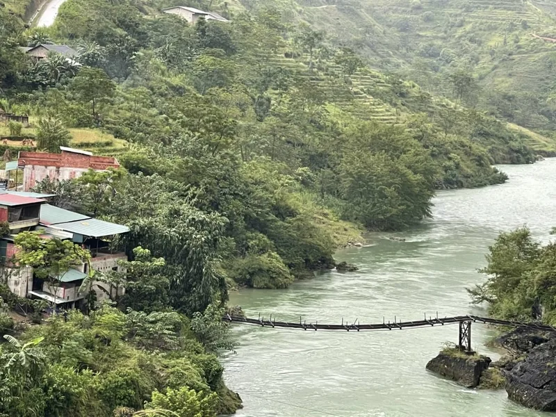 A suspension bridge crosses the turquoise Nho Que River beneath green terraced hills in Ha Giang.