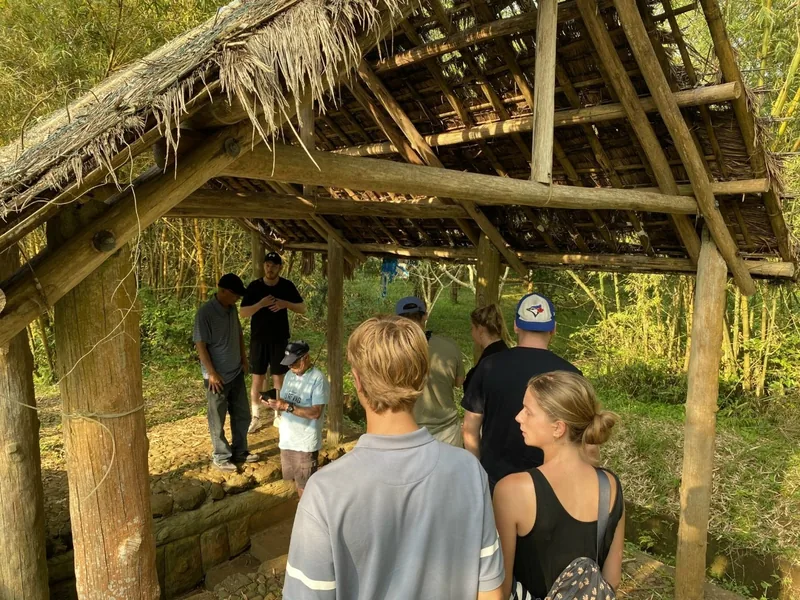 Tourists gather under a thatched-roof shelter while a guide explains a former tunnel site on the Hue DMZ tour.