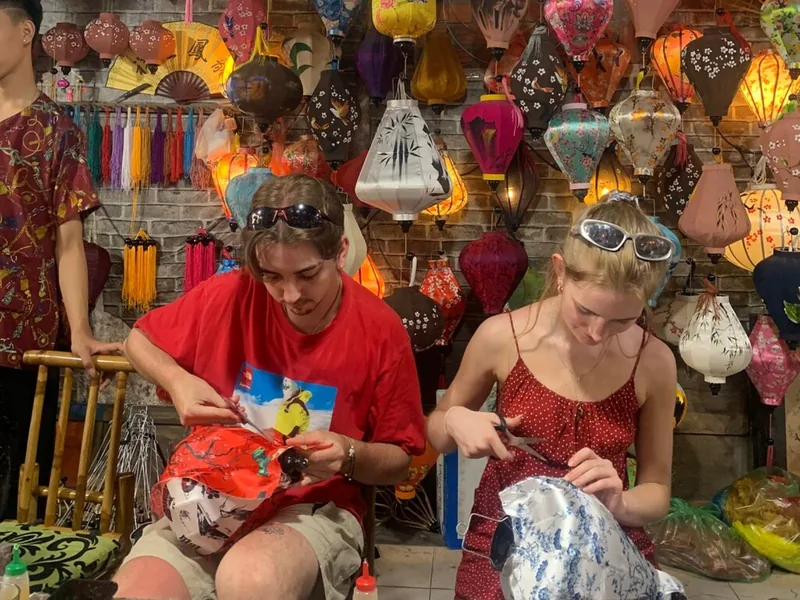 Tourists try making colorful silk lanterns in a shop in Hoi An, surrounded by hanging lanterns.