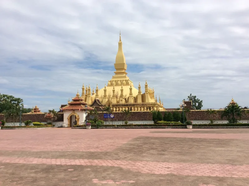 The golden spire of Pha That Luang stupa rises above its walled courtyard in Vientiane under a cloudy sky.