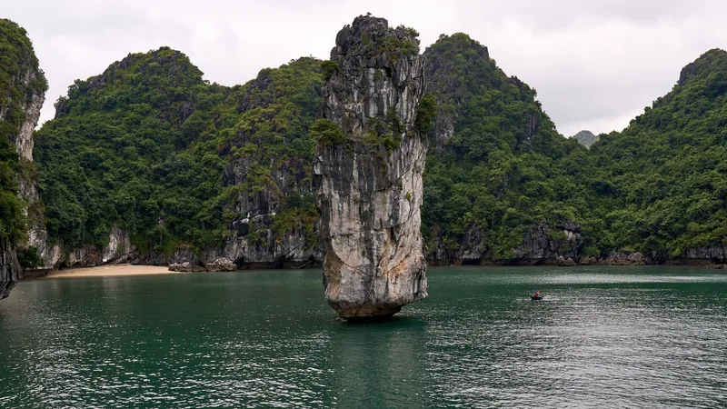 A tall limestone pillar rises from the emerald waters of Ha Long Bay, flanked by karst islets and a small beach.