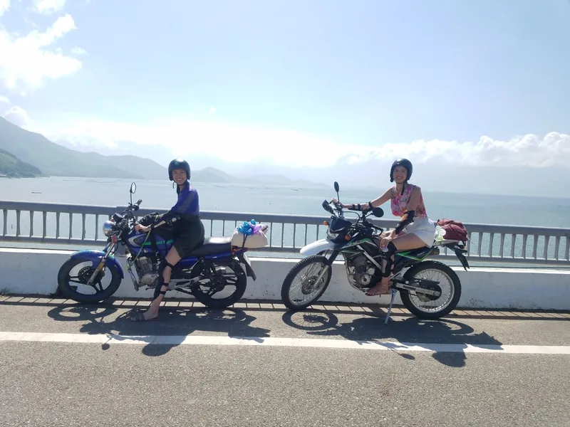 Two women pause on motorbikes atop the Hai Van Pass road between Hue and Hoi An, mountains and sea behind them.