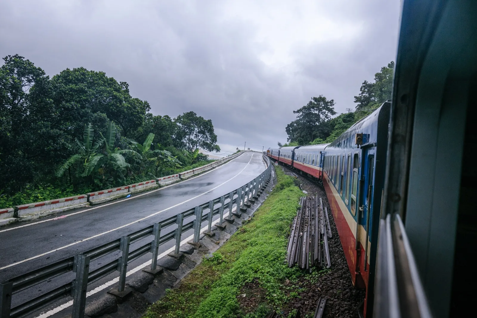 A train winds along tracks beside a wet coastal road through green hills near Da Nang on an overcast day.
