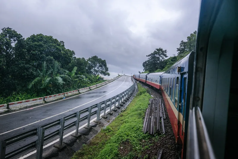 A train winds along tracks beside a wet coastal road through green hills near Da Nang on an overcast day.