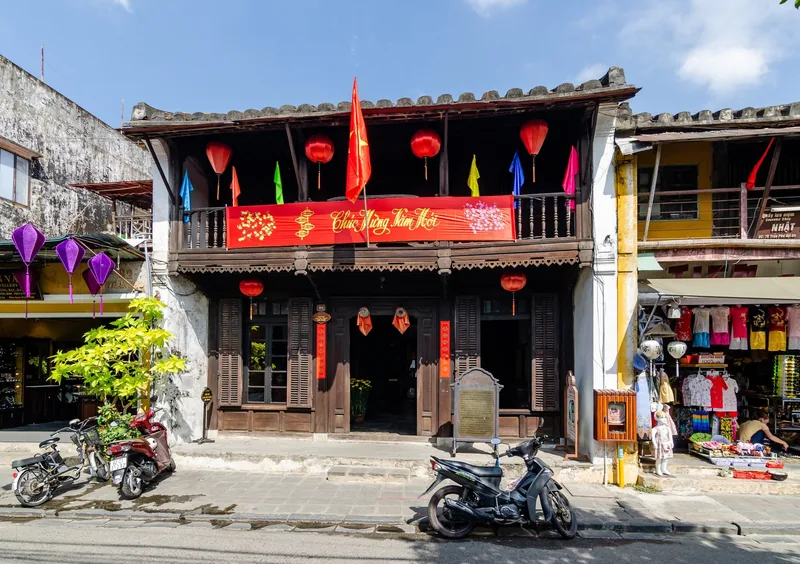 A traditional wooden two-story house in Hoi An decorated with red lanterns and flags, with motorbikes parked outside.