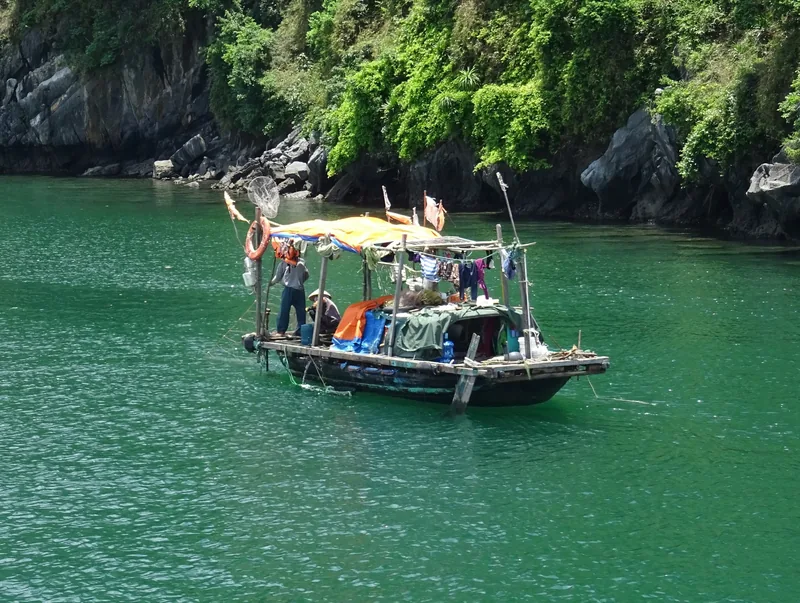 A small fishing boat with a canvas awning drifts on the emerald water of Ha Long Bay beneath limestone cliffs.