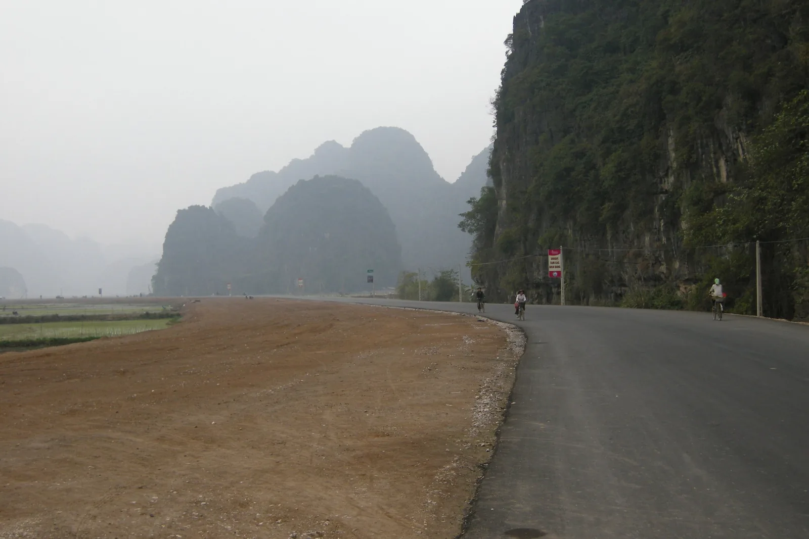 Cyclists ride a quiet road beneath hazy limestone karsts near rice fields in Ninh Binh.