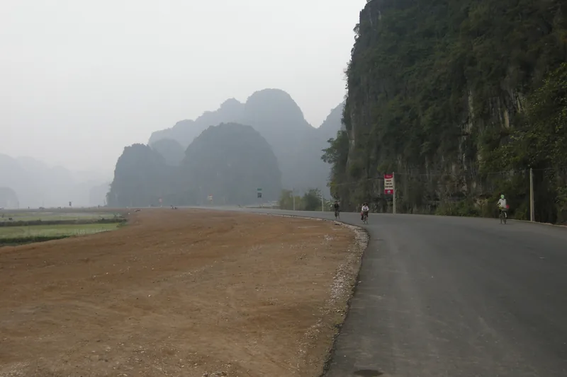 Cyclists ride a quiet road beneath hazy limestone karsts near rice fields in Ninh Binh.