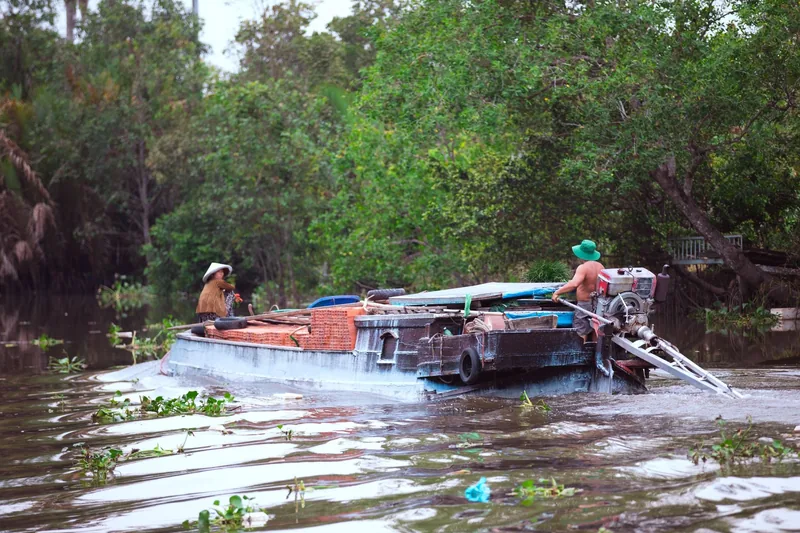 A cargo boat carrying bricks navigates a lush green river channel in the Mekong Delta.