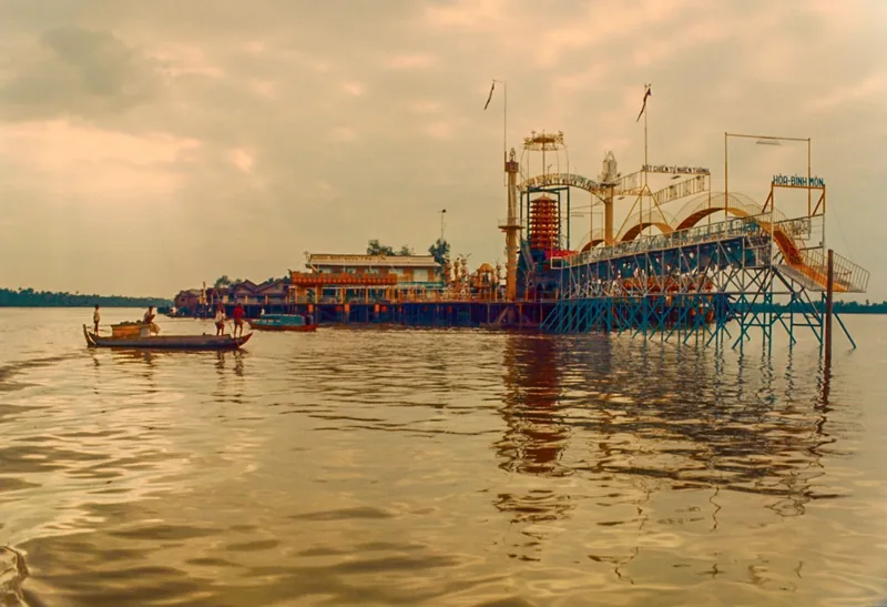A small wooden boat passes a floating structure with towers and archways on the Mekong River, likely the Coconut Monk's Dao.