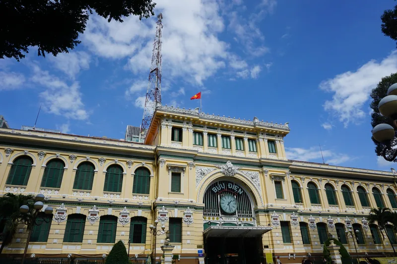 The colonial-era Saigon Central Post Office facade with arched windows and a Vietnamese flag under a blue sky.