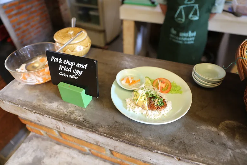 A plate of com tam pork chop and fried egg with rice sits on a wooden counter beside a chalkboard sign in Vietnam.