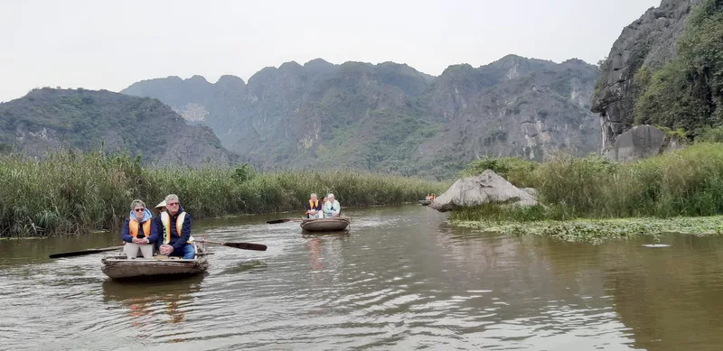 Tourists in small rowboats glide along the river at Van Long Nature Reserve, surrounded by limestone karst mountains.