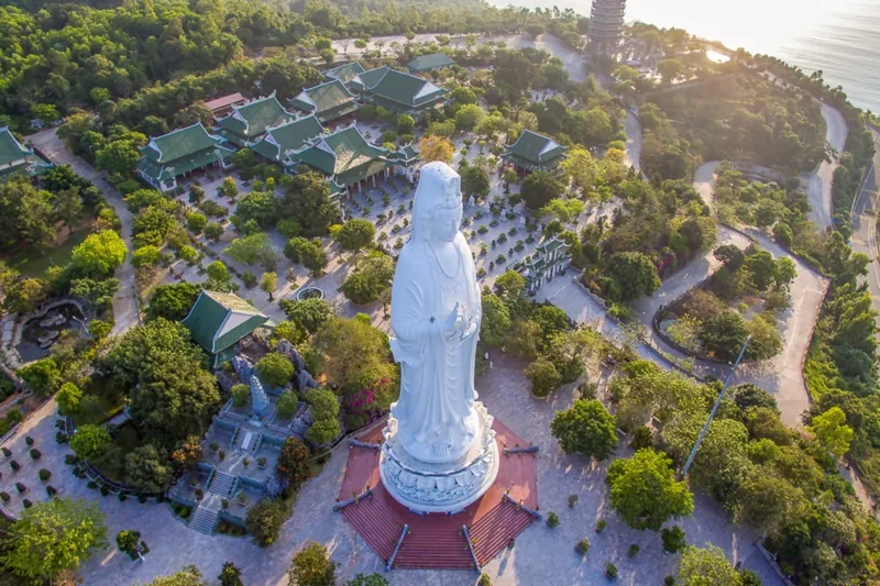 Aerial view of the tall white Lady Buddha statue rising above Linh Ung Pagoda's green-roofed temple complex near the coast.