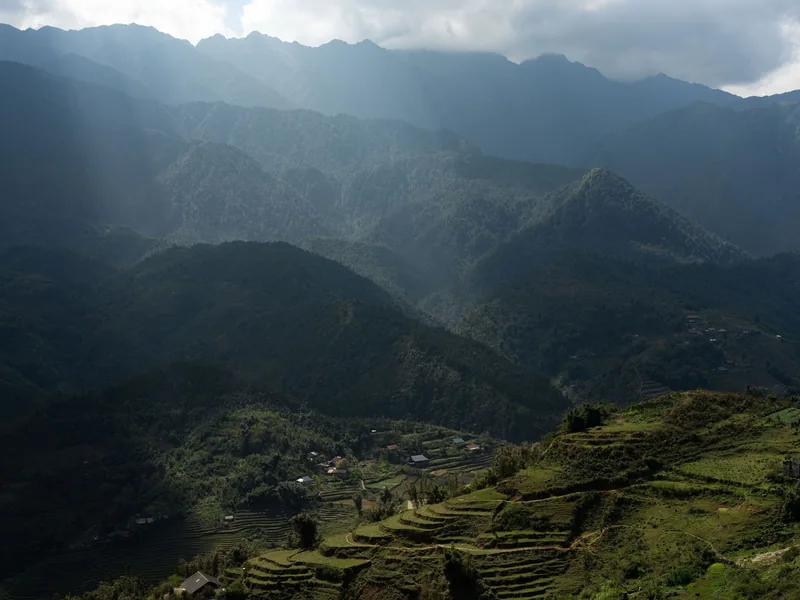 Sunbeams pierce clouds over layered mountain ridges and terraced rice fields near Sapa.