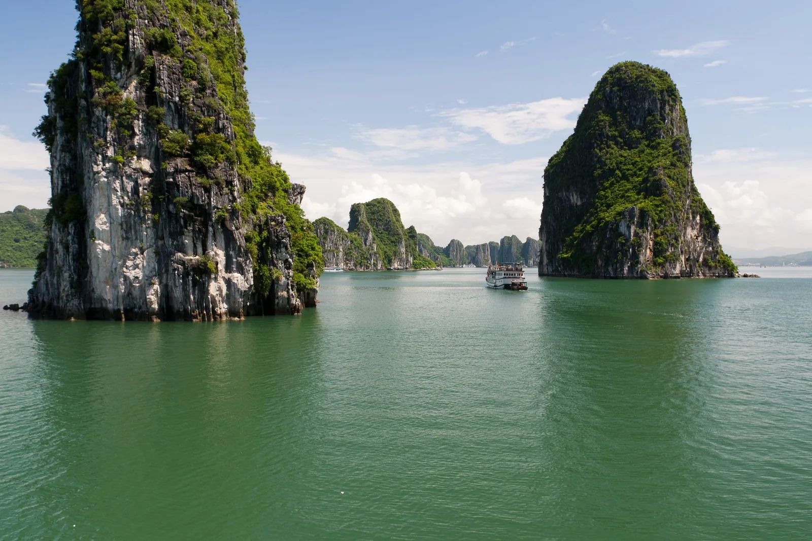 Limestone karsts rise from emerald water as a small cruise boat sails through Ha Long Bay.