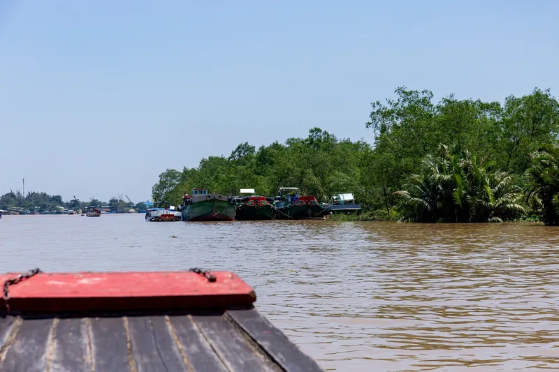 Cargo barges moored along a muddy river lined with palms, viewed from a wooden boat deck in the Mekong Delta.