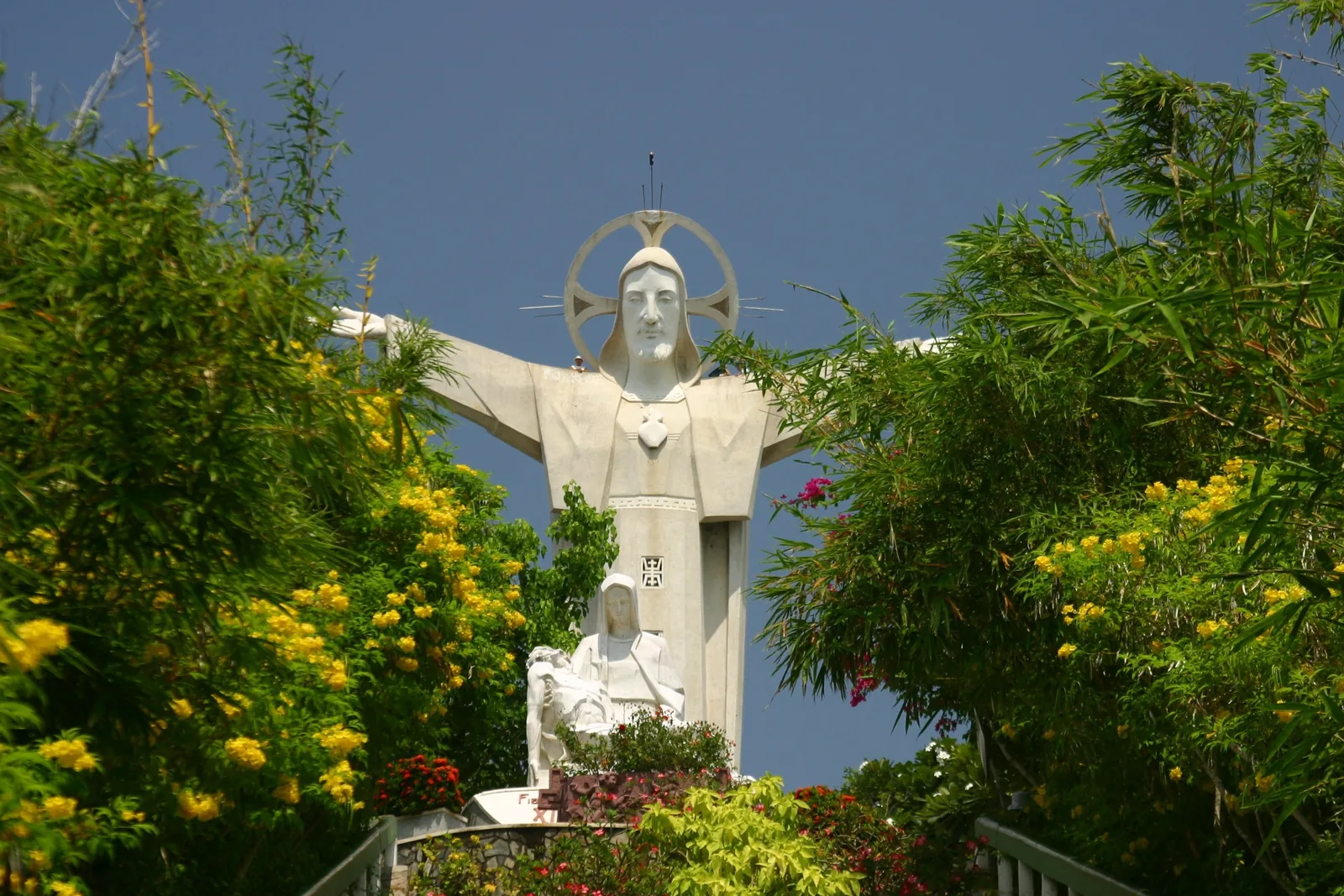 The Christ of Vung Tau statue with outstretched arms rises above lush green trees and yellow flowers.