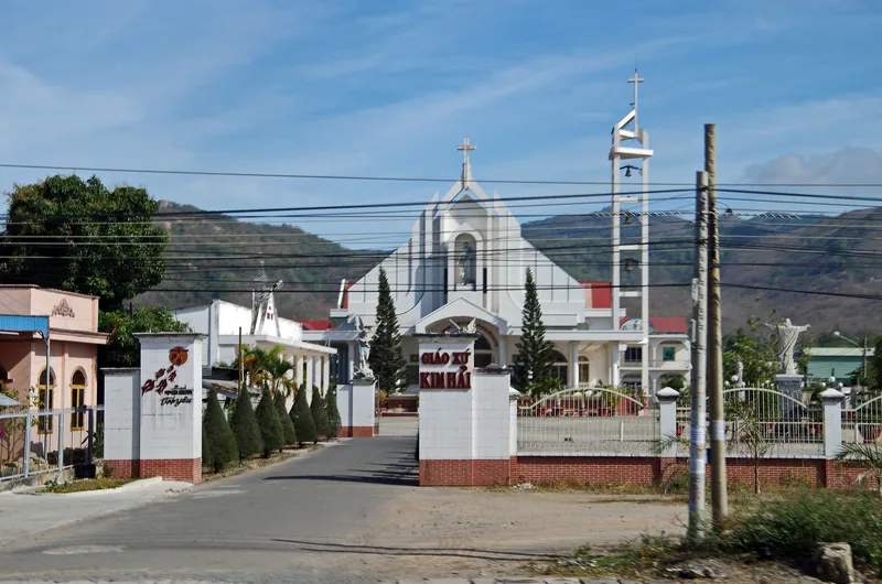 White Catholic church with bell tower stands behind a gated courtyard near Vung Tau, hills in the background.